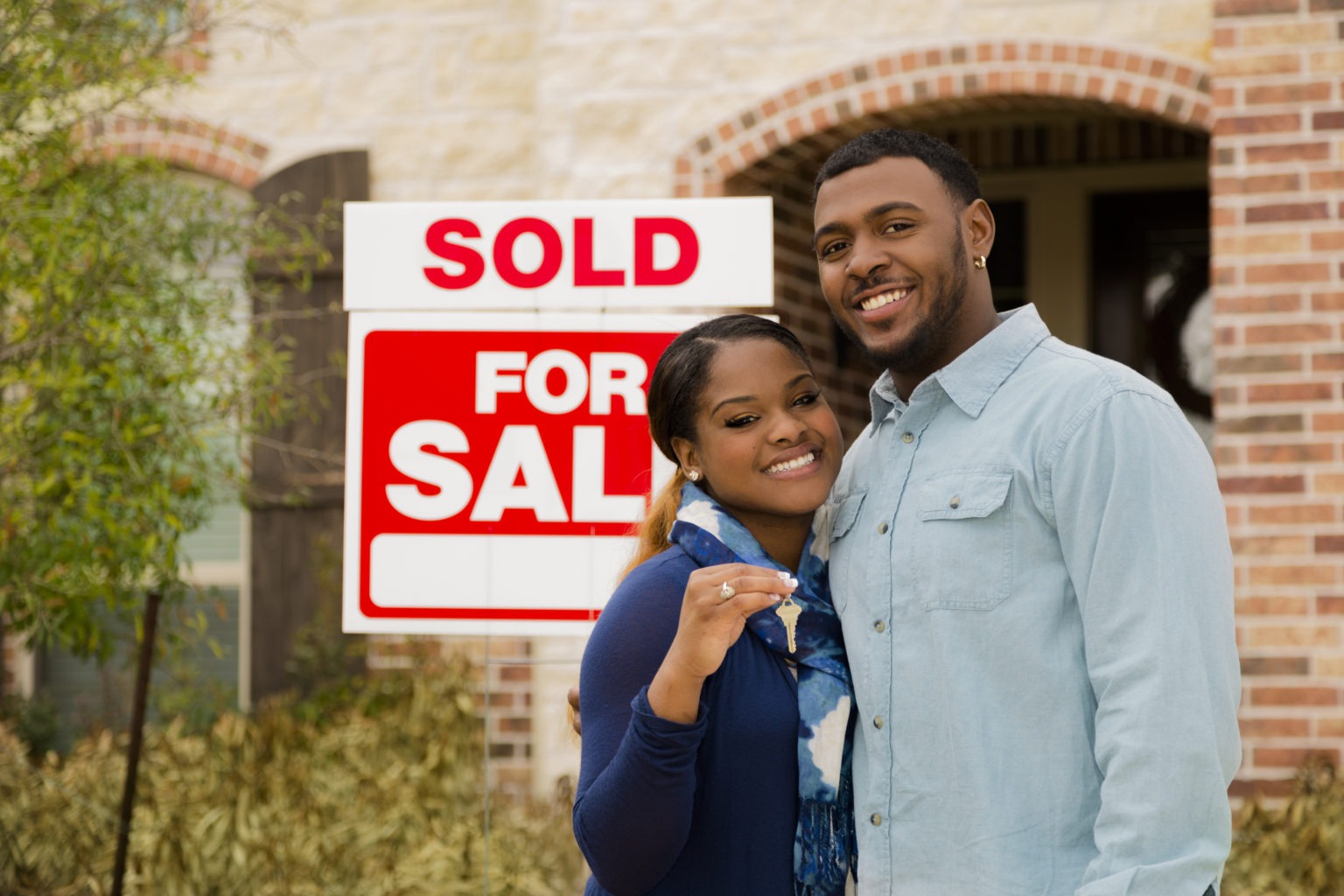 African descent couple shows off key after purchasing a new home. 'Sold',  'For Sale' sign in background. Front entrance of this beautiful brick and stone home.